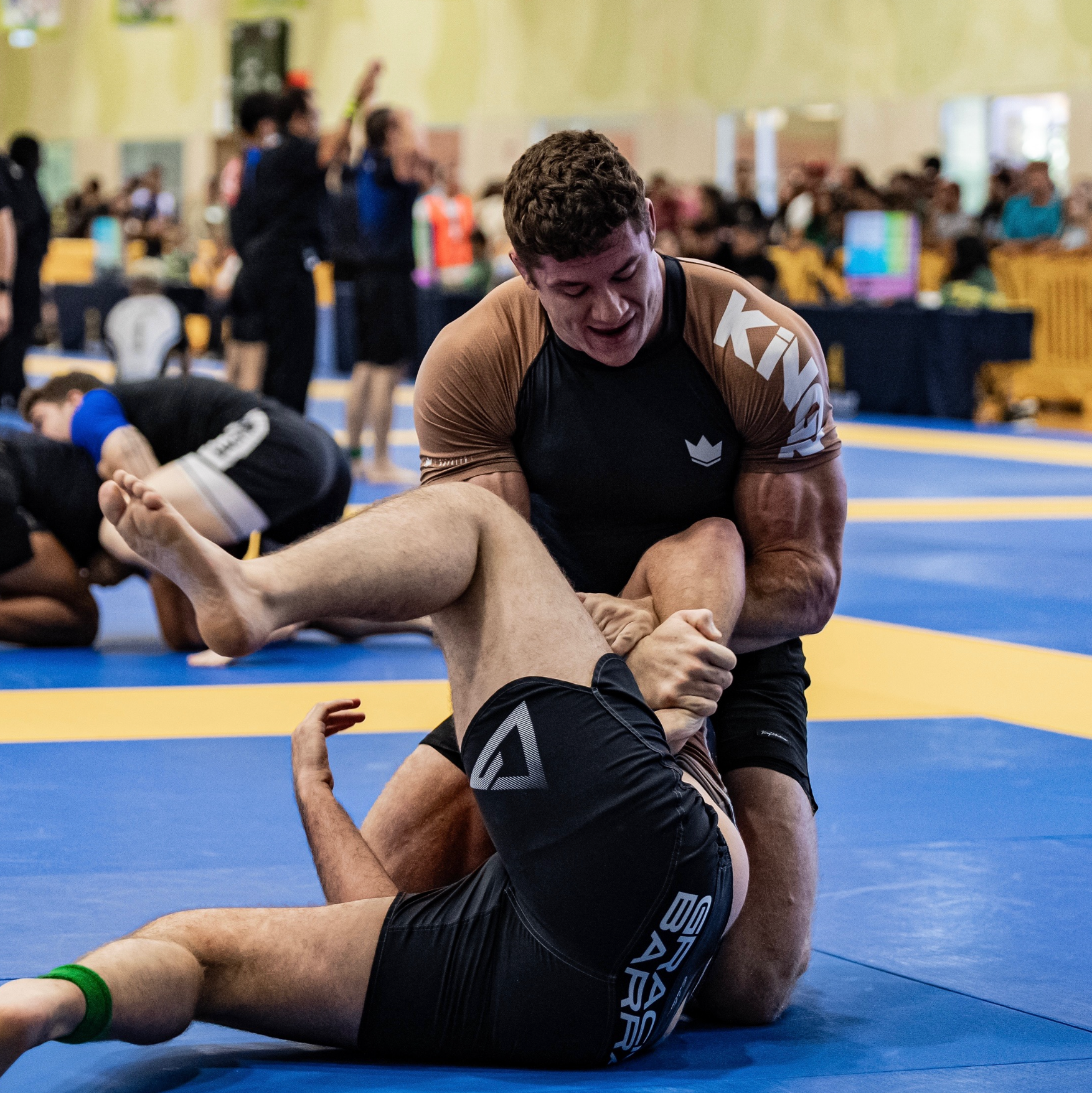 Two individuals engaged in a grappling match on a blue mat with spectators in the background.
